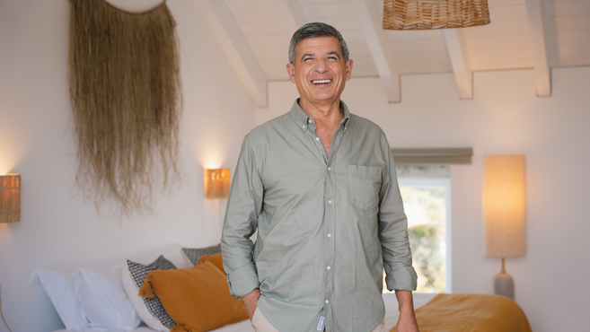 Zelfverzekerde, glimlachende man in een groen shirt, staand in een moderne en sfeervol verlichte slaapkamer met warme kleuren, een wanddecoratie van gevlochten materiaal en mosterdgeel beddengoed.
