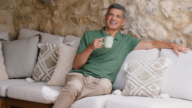 Een lachende man zit comfortabel op een buitenbank met witte kussens en houdt een keramische mok vast. Hij draagt een groen poloshirt en een beige broek en leunt tegen een stenen muur.