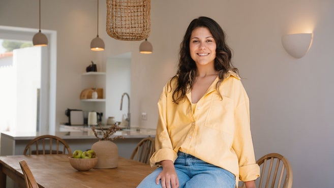 Een lachende jonge vrouw in een geel overhemd en blauwe jeans zit op een houten eettafel in een moderne, open keuken en eetkamer.