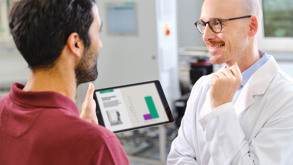 Twee mensen, een nefroloog en een technicus, in gesprek in een waterbehandelingsruimte. De nefroloog draagt een witte laboratoriumjas en de andere houdt een tablet vast waarop het verbruik van hulpbronnen bij dialyse wordt weergegeven.