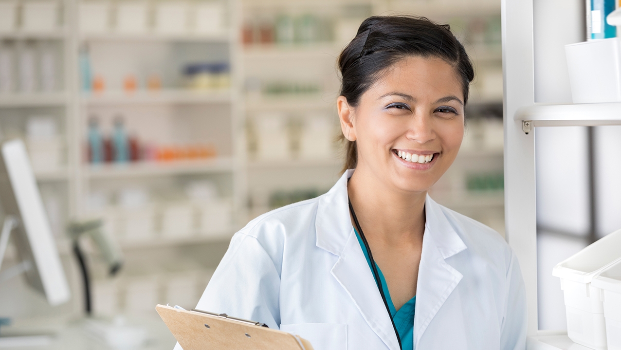 Smiling mid adult Asian female pharmacist stands in her pharmacy. She is smiling at the camera and is holding a clipboard.
