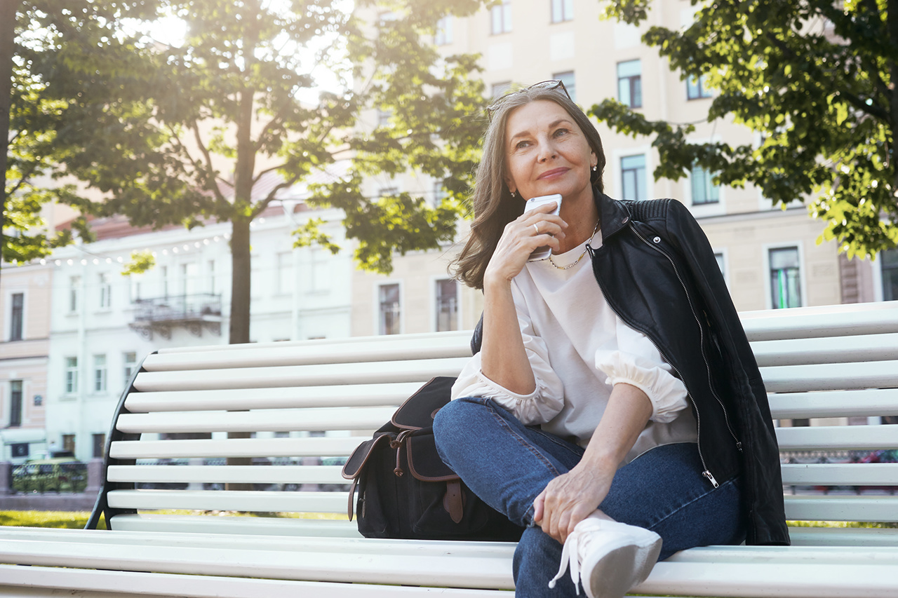 Low angle view of positive stylish middle aged female in trendy leather jacket, blue jeans and sneakers having pensive look, sitting comfortably on bench with cell phone, thinking where to have lunch