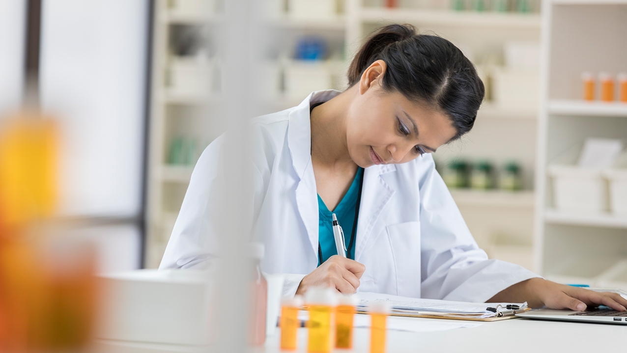 Mid adult female pharmacist concentrates while writing something on a clipboard. She is also using a laptop.