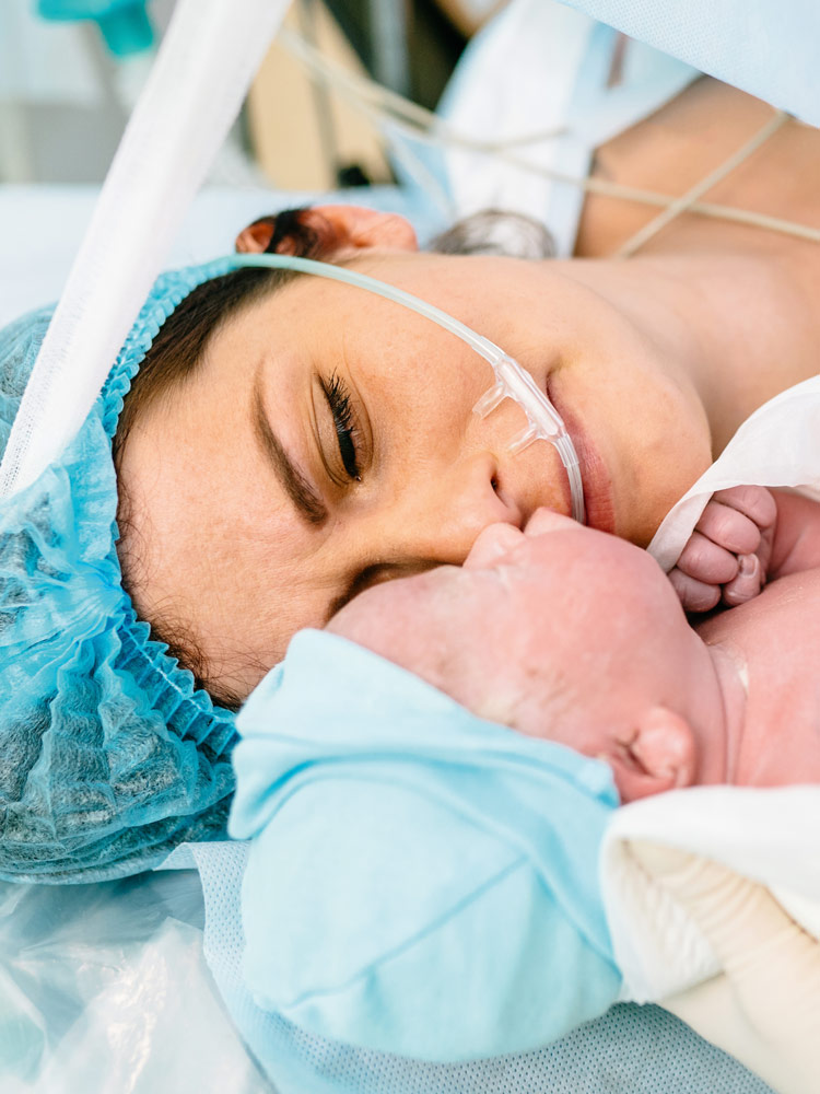 Een moeder ligt na een keizersnede in de operatiekamer en kijkt naar haar pasgeboren baby, die een verpleegkundige haar laat zien. Ze ziet er vermoeid uit, maar ook opgelucht. Ze draagt een operatiemuts over haar donkere haar, de pasgeborene is in een witte doek gewikkeld en draagt een lichtblauwe muts.