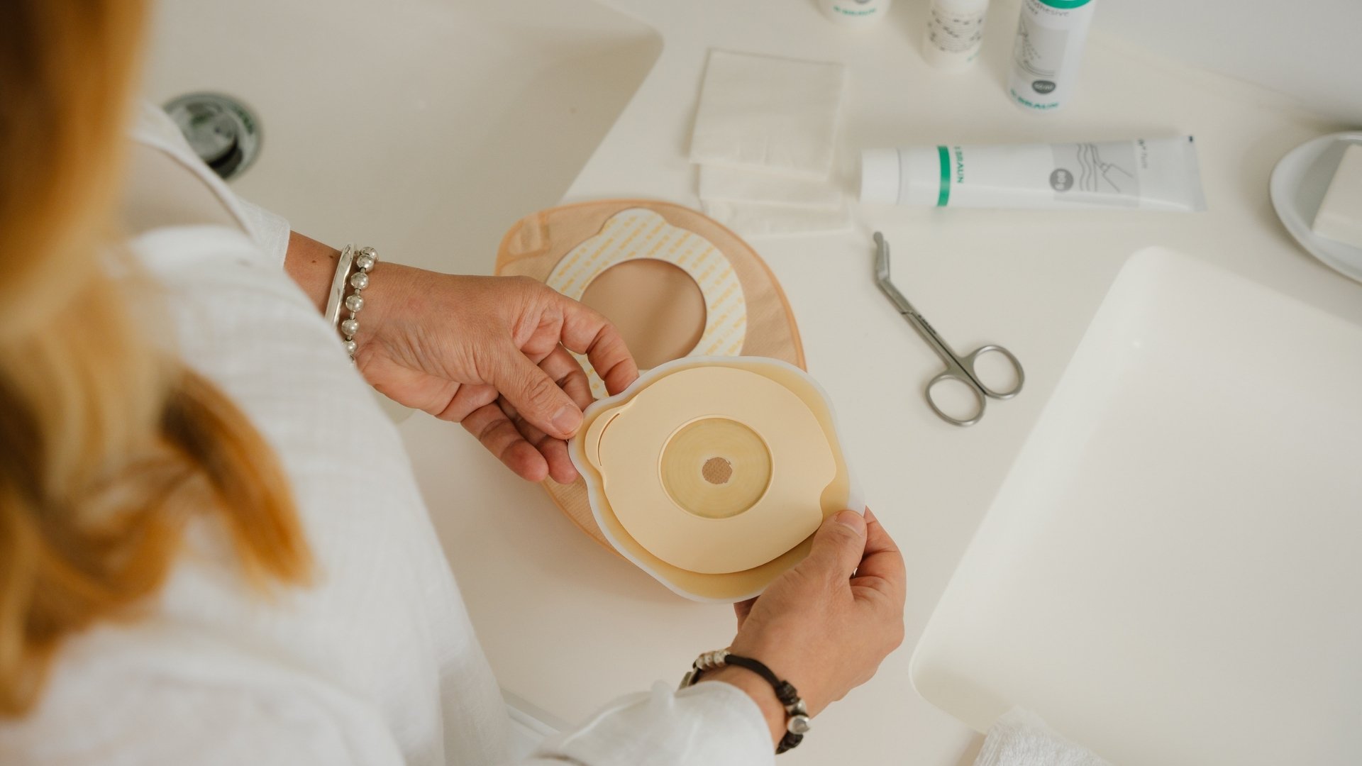 Een bovenaanzicht van een persoon die een stomaplak vasthoudt boven een witte badkamertafel. Op de achtergrond een beige stomazak, stomaschaar en stoma-accessoires.